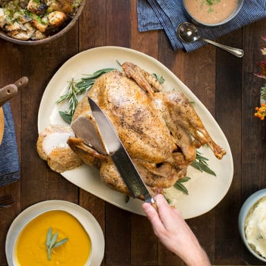 A hand about to cut a whole turkey on a tablescape of butternut squash soup, mashed potatoes, salad