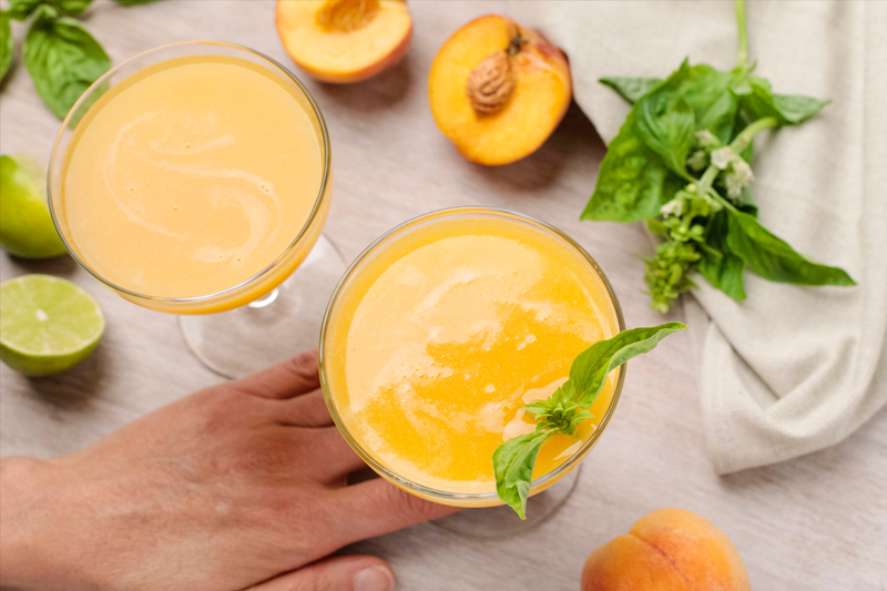 Hand holding coupe glass of blended peach mojito with surface covered with limes, peaches and basil leaves