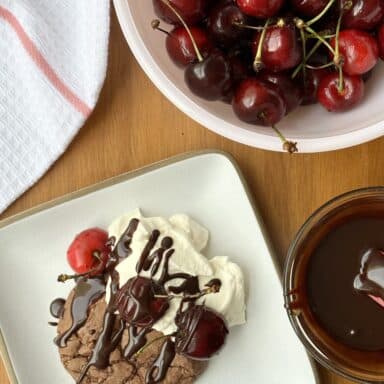 A chocolate cookie on a white plate, drizzled with chocolate sauce, with a dollop of whipped cream, and topped with fresh cherries.