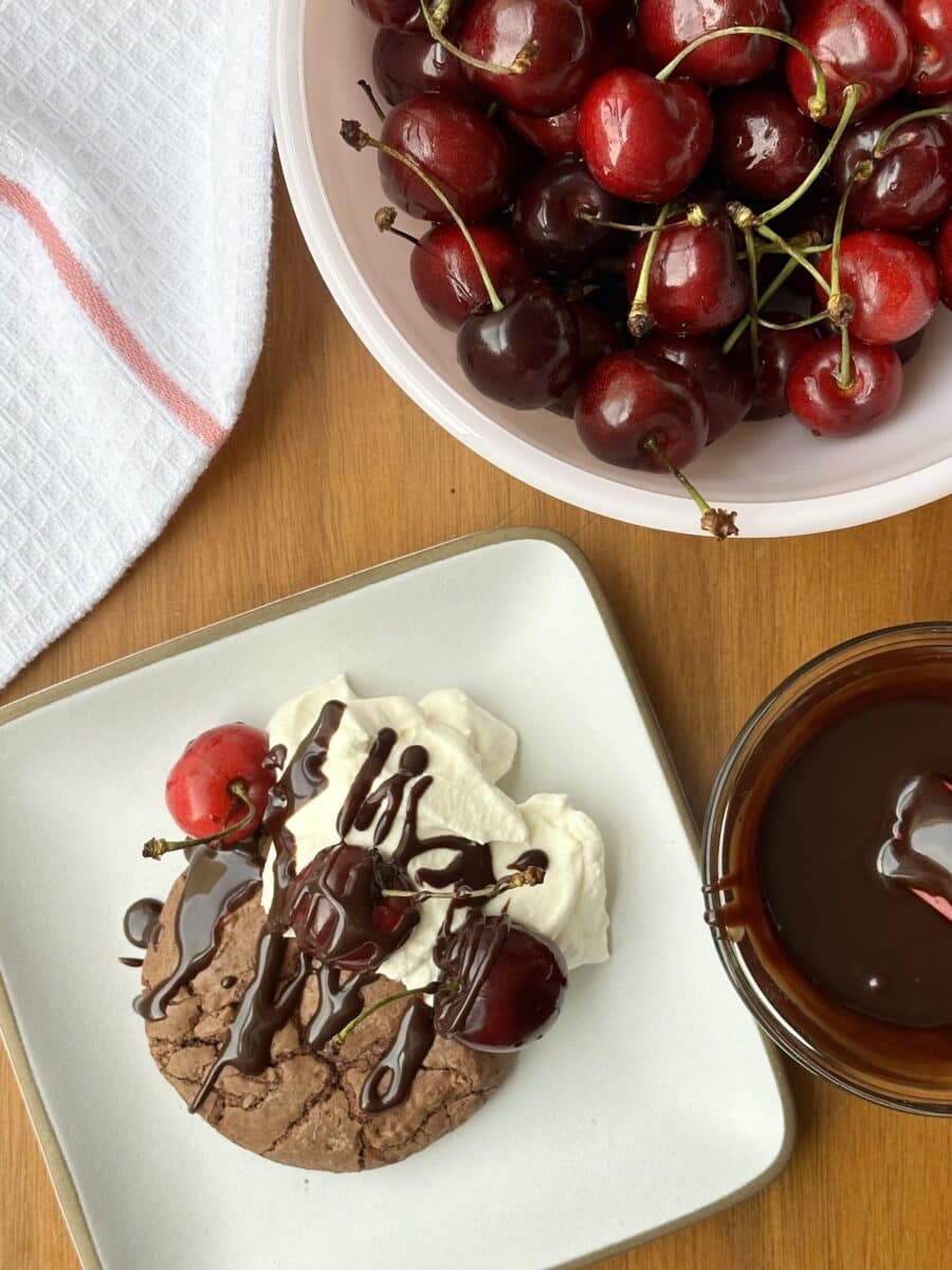 A chocolate cookie on a white plate, drizzled with chocolate sauce, with a dollop of whipped cream, and topped with fresh cherries.