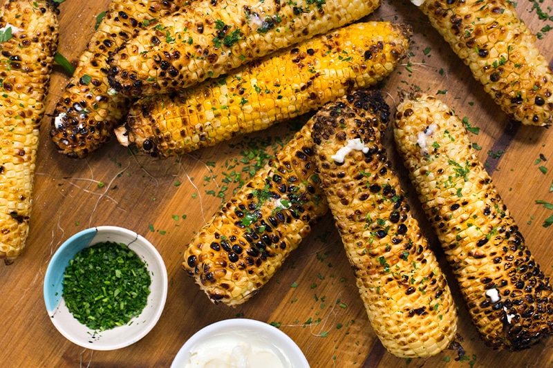 Eight ears of corn on a wooden surface, grilled and covered with herbs with herbs and cream on the side
