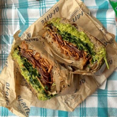 The Bernal Heights signature sandwich from Bi-Rite Market, cut in half, loaded with bacon, avocado, and crispy sweet potatoes on parchment paper and a checkered tablecloth.