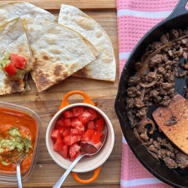 Quesadillas being served with tomatoes and guacamole on a wooden cutting board, next to a cast iron skillet with ground beef.