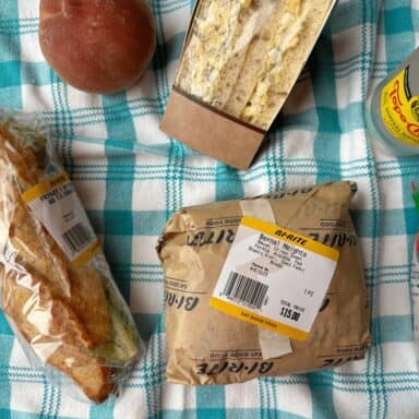 Three sandwiches in wrappers, a peach, a bottle of sparkling water, and a bag of chips on a checkered tablecloth.
