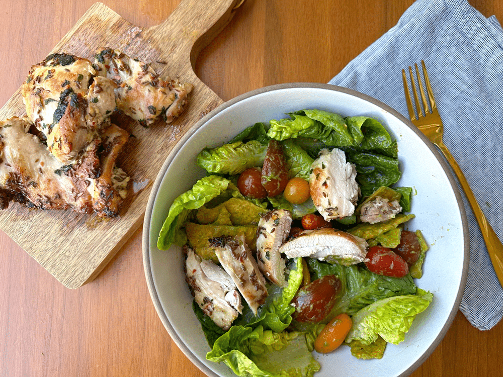 Herbed chicken thighs on a wooden cutting board, next to a bowl of salad with sliced chicken on top.