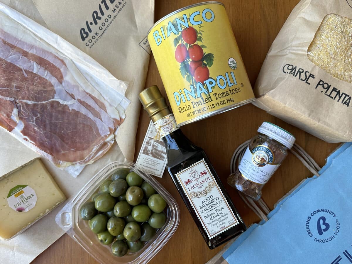 Unpacking a Bi-Rite grocery bag, with packages of prosciutto, tomatoes, polenta, cheese, olives, vinegar, and anchovies spilling out on a table.