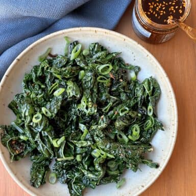 A Shallow Bowl Filled With Sautéed Kale Leaves, With Spoon Digging Into A Jar Of Bright Red Chili Crisp
