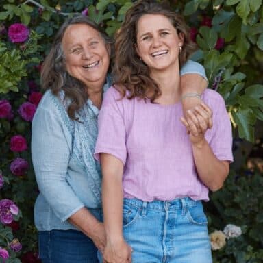 Laughing Dru Rivers And Hannah Muller Of Full Belly Farm, Facing The Camera, Dru's Arm Draped Across Hannah Amongst A Lush Canopy Of Flowers