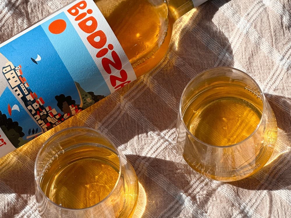 A Bottle Of Biddizza Laid Down On A Picnic Blanket Next To Two Full Glasses