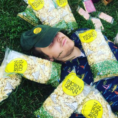 Bjorn Qorn Employee Wearing Brand Cap Surrounded By Bags Of Popcorn On Grass