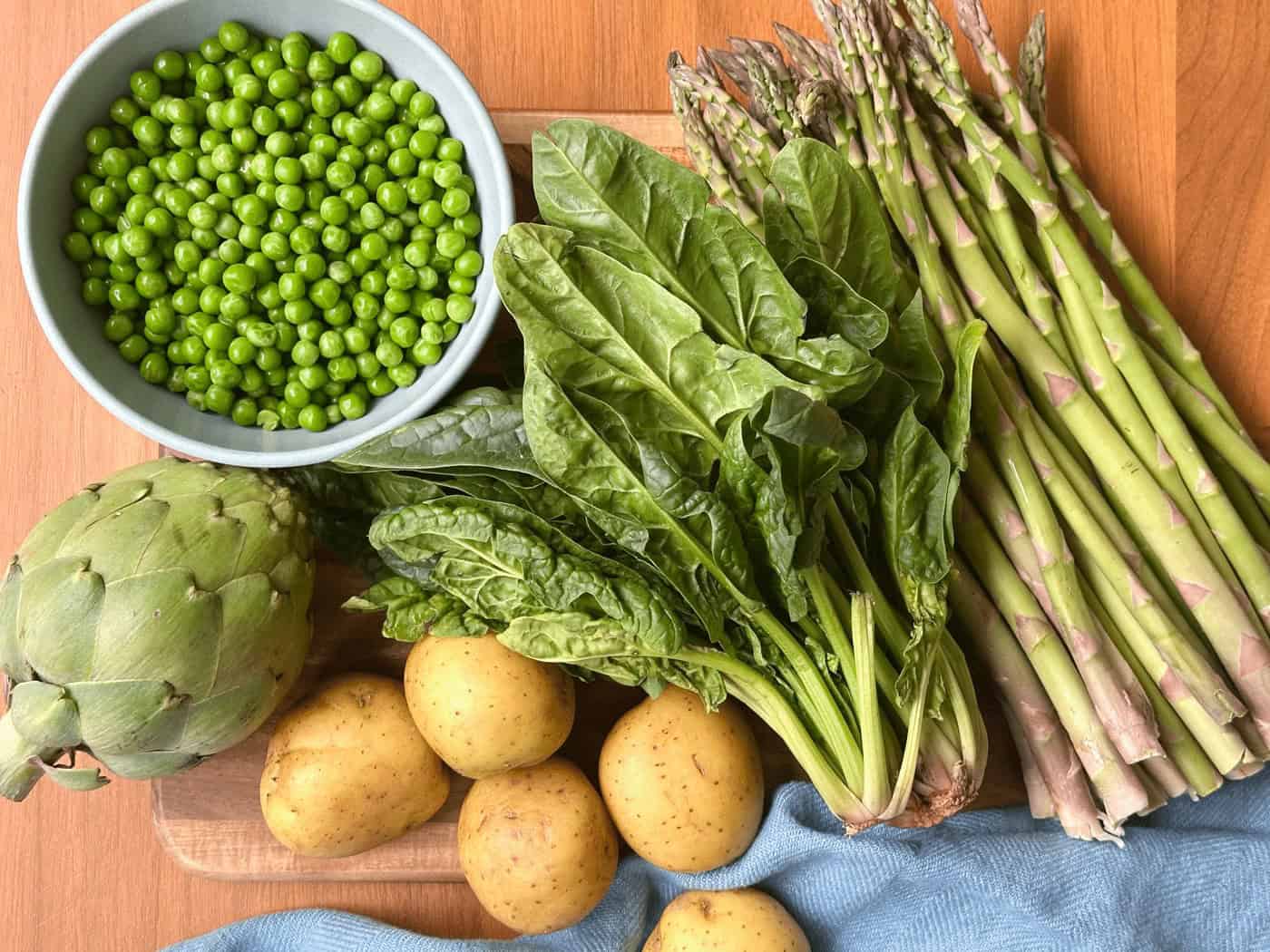 Mis En Place Of Bowl Of Peas Artichoke Golden Potatoes Bunch Of Lettuce Bunch Of Asparagus On Wood Cutting Board With Blue Napkin