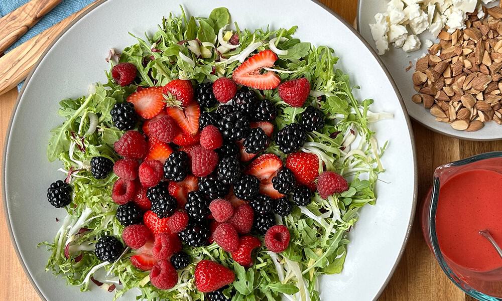 Summer Berry Salad Plated With Frisee And Chese And Almonds And Dressing On The Right Side With Blue Napkin And Tongs